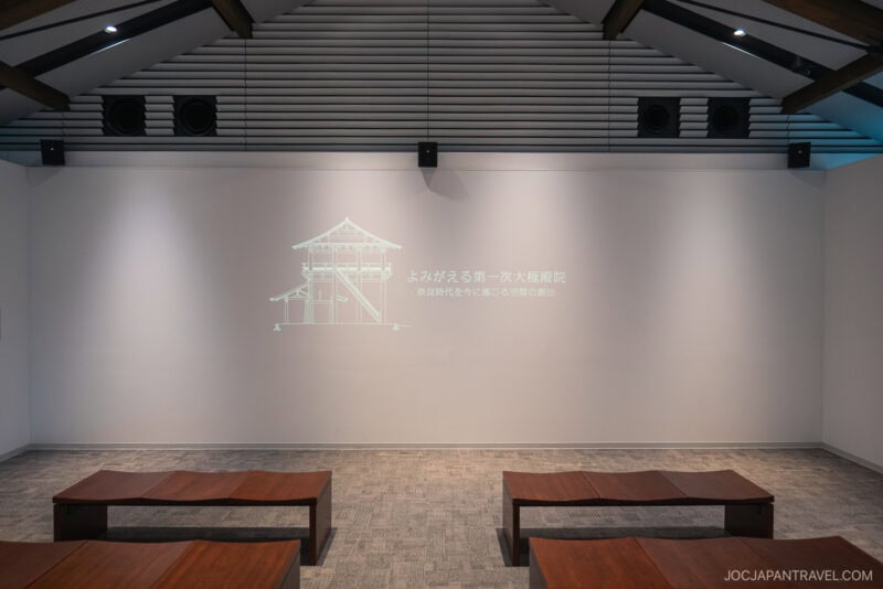A small theater room at the Nara Palace Site features wooden benches facing a white wall, where a faint architectural drawing and Japanese text are projected. The space boasts a modern ceiling with visible beams and speakers.