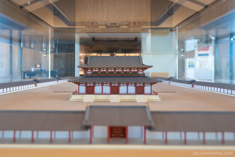A detailed scale model of a traditional Japanese palace, inspired by the Nara Palace Site, is displayed in a glass case with modern architecture reflected in the background. The structure features red columns, tiered roofs, and a symmetrical layout.