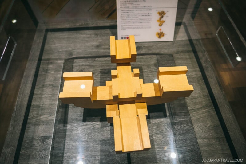 A wooden puzzle or sculpture with interlocking rectangular pieces is displayed in a glass case on a dark wood surface at the Nara Palace Site, with an informational sign in the background.