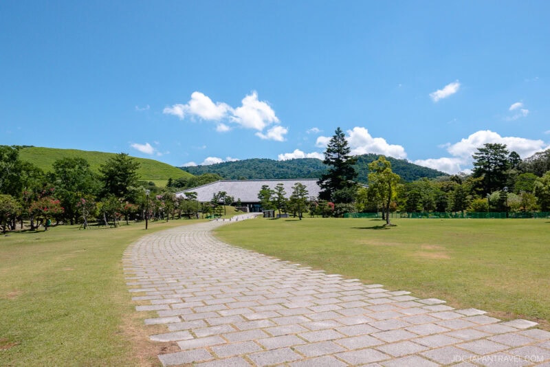 A stone pathway curves through wide, grassy Nara Park with scattered trees, leading toward a building. Green hills and a blue sky with scattered clouds frame the peaceful scene.