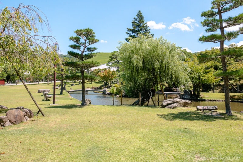 A serene Japanese garden inspired by Nara Park features a grassy lawn, various trees, a small stream, rocks, and benches under a bright blue sky with scattered clouds.