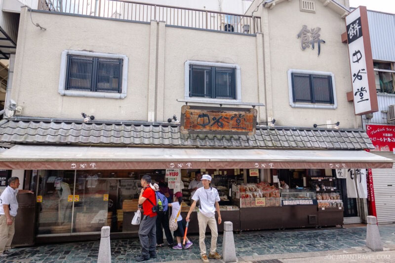 A traditional Japanese shop in Nara Naramachi with a tiled roof, signs in Japanese, and people browsing outside. A man and child hold hands at the entrance, while others admire goods displayed along Mochiidono Street.