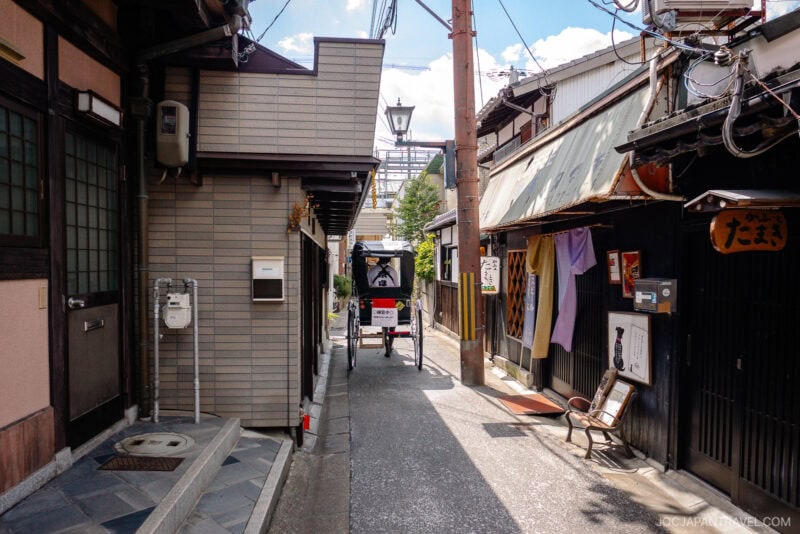 A narrow street in Nara’s Naramachi district features traditional wooden buildings and a rickshaw being pulled by a person. Signs, lanterns, and power lines hang above under the bright blue Mochiidono sky with clouds.