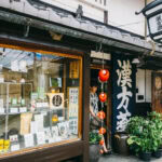Naramachi and Mochiidono Center Gai – Exploring Nara’s Historic Heart and Lively Shopping Street A traditional Japanese shop in Nara Naramachi Mochiidono, with a wooden exterior, paper lanterns, large kanji sign, and shelves displaying products in the window. A person stands in the doorway next to green potted plants.