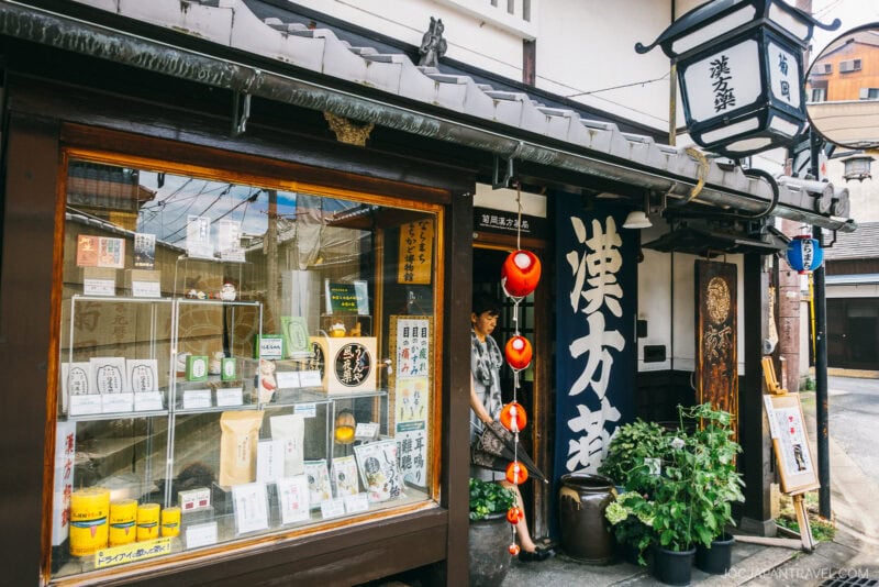 A traditional Japanese shop in Nara Naramachi Mochiidono, with a wooden exterior, paper lanterns, large kanji sign, and shelves displaying products in the window. A person stands in the doorway next to green potted plants.