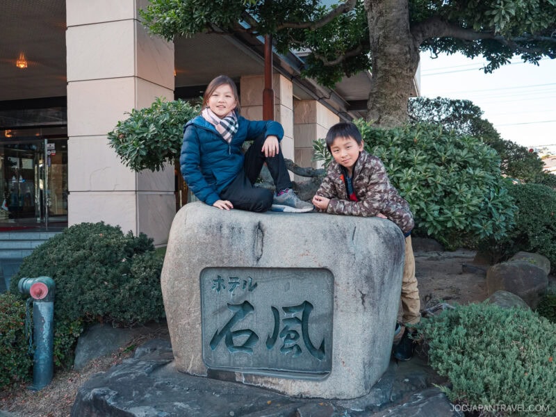 Two children sit and pose on a large stone sign with Japanese writing at Isawa Onsen Yamanashi, in front of a building surrounded by greenery. Both are smiling and dressed in winter clothing.