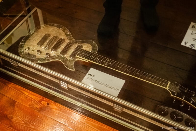 An electric guitar with a shiny, reflective body is displayed in a glass case on a wooden floor at Isawa Onsen Yamanashi, accompanied by a descriptive placard and some visible feet in the background.