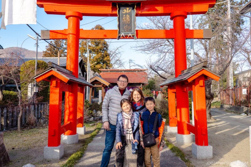 A family of four smiles while posing under a bright red torii gate at a shrine in Isawa Onsen Yamanashi on a sunny day. Trees and traditional buildings are visible in the background.