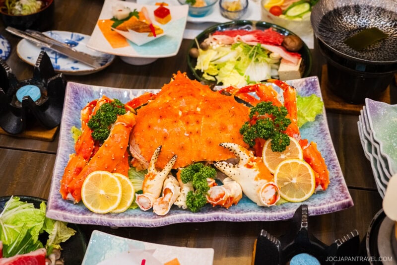 A large platter features a whole cooked crab surrounded by crab legs, garnished with lemon slices, lettuce, and parsley. The table is set with various Japanese dishes and utensils, evoking the culinary delights of Isawa Onsen Yamanashi.