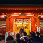 Ringing in the New Year at Isawa Onsen (石和温泉), Yamanashi People stand in line at night in front of a brightly lit Shinto shrine with red pillars and traditional decorations in Isawa Onsen Yamanashi, preparing to pray or make offerings.
