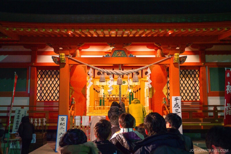 People stand in line at night in front of a brightly lit Shinto shrine with red pillars and traditional decorations in Isawa Onsen Yamanashi, preparing to pray or make offerings.