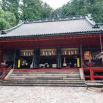Nikko Futarasan Jinja (日光二荒山神社) – A Shrine of Mountains and Ancient Cedars Traditional Japanese temple with a sloped roof, red pillars, stone steps, and two stone lanterns at the entrance, surrounded by lush green trees—evoking the serene beauty of Nikko Futarasan Jinja.