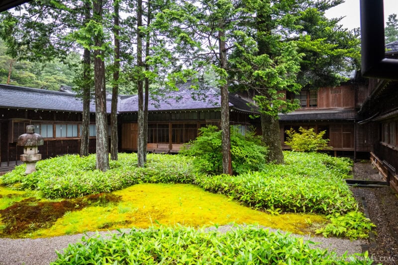 A traditional Japanese courtyard garden with moss, green shrubs, and tall trees, surrounded by dark wooden buildings and a stone lantern on the left side—evoking the serene beauty of Nikko Tamozawa Imperial Villa Memorial Park.