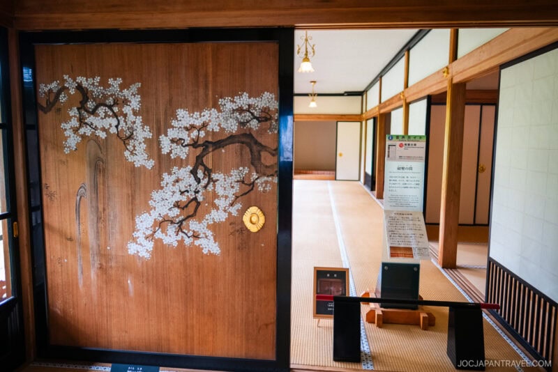 A traditional Japanese room with tatami mats and sliding wooden doors, one featuring a painted cherry blossom tree, reminiscent of the serene interiors found at Nikko Tamozawa Imperial Villa Memorial Park. The space features display signs and soft natural lighting.