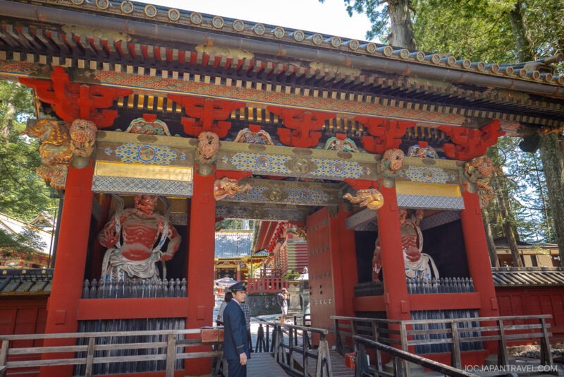 A person stands near the entrance of the colorful, ornate Nikko Toshogu Shrine gate adorned with detailed carvings, red pillars, and fierce guardian statues, surrounded by trees.