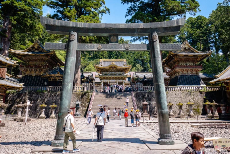 Visitors walk through a large torii gate toward ornate temple buildings at Nikko Toshogu Shrine, surrounded by tall trees at this historic site in Japan, with steps leading up to the main shrine.