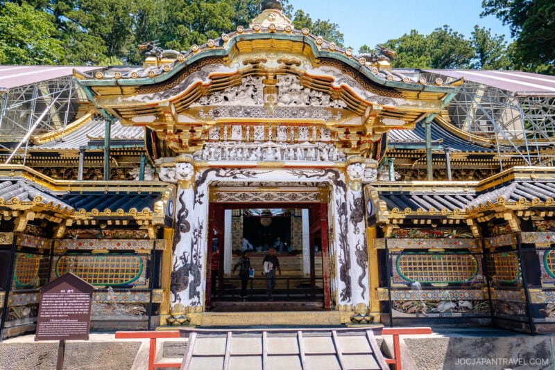 Ornate, colorful gate with intricate carvings and gold accents at Nikko Toshogu Shrine in Japan, surrounded by trees, with visitors visible inside the open entrance.