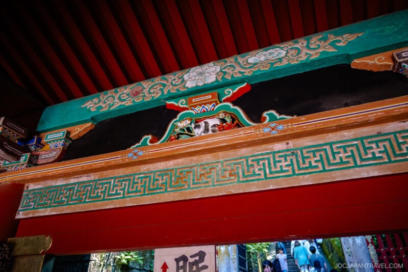 Decorative details of a traditional Japanese temple gate at Nikko Toshogu Shrine, featuring colorful painted patterns, intricate floral designs, and woodwork. People can be seen walking through the gate in the background.