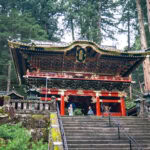 Nikkosan Rinnoji Taiyuin (日光山輪王寺大猷院) – The Resting Place of Tokugawa Iemitsu A colorful traditional Japanese temple, Nikkosan Rinnoji Taiyuin, with intricate carvings and gold details stands among tall trees. Several visitors are on the stone steps leading up to the entrance.