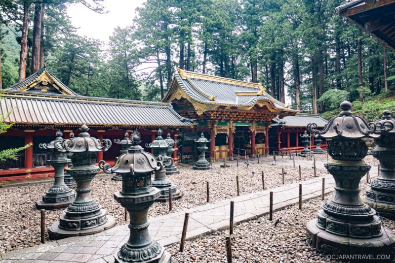 Traditional Japanese temple, Nikkosan Rinnoji Taiyuin, with ornate golden roof details, surrounded by tall trees and stone lanterns on a gravel courtyard. Red wooden structures and the green forest create a serene, historic atmosphere.