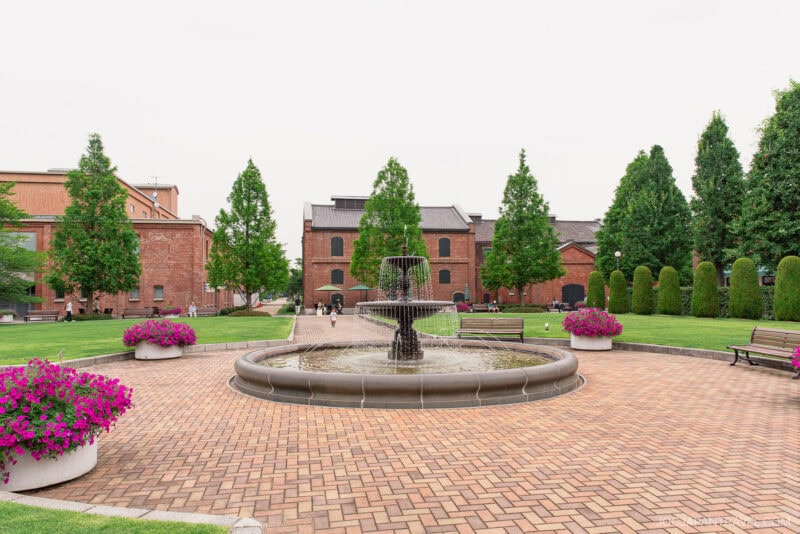 A circular fountain surrounded by brick pavement sits in the Noritake Garden courtyard, with benches, large flower pots filled with pink flowers, green trees, and red brick buildings in the background.