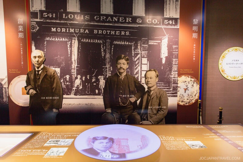 Historical exhibit display at Noritake Garden featuring three men in front of a vintage Morimura Brothers storefront photo, with informational panels and a round interactive display table in the foreground.