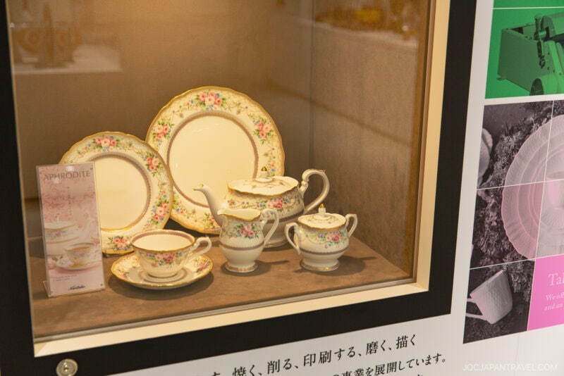 A display case showcases an elegant, floral-patterned Noritake Garden porcelain tea set with teapot, sugar bowl, creamer, cups, saucers, and plates. A card labeled “APHRODITE” stands beside the set.