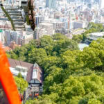 Kobe Nunobiki Ropeway and Herb Gardens (神戸布引ロープウェイ・ハーブ園) Aerial view from a red cable car on the Nunobiki Ropeway descending toward a station surrounded by green trees and the Herb Gardens, with a cityscape of tall buildings and streets in the background.