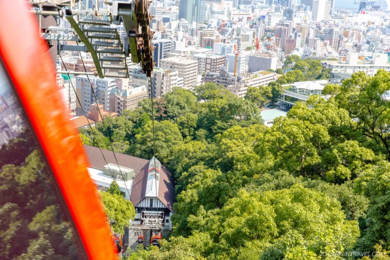 Aerial view from a red cable car on the Nunobiki Ropeway descending toward a station surrounded by green trees and the Herb Gardens, with a cityscape of tall buildings and streets in the background.