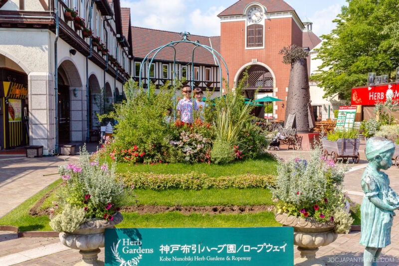 A lush garden display with colorful flowers and greenery sits before shops at Kobe Nunobiki Ropeway and Herb Gardens in Japan. A man stands in a green gazebo, while a statue of a girl and a green sign are visible nearby.