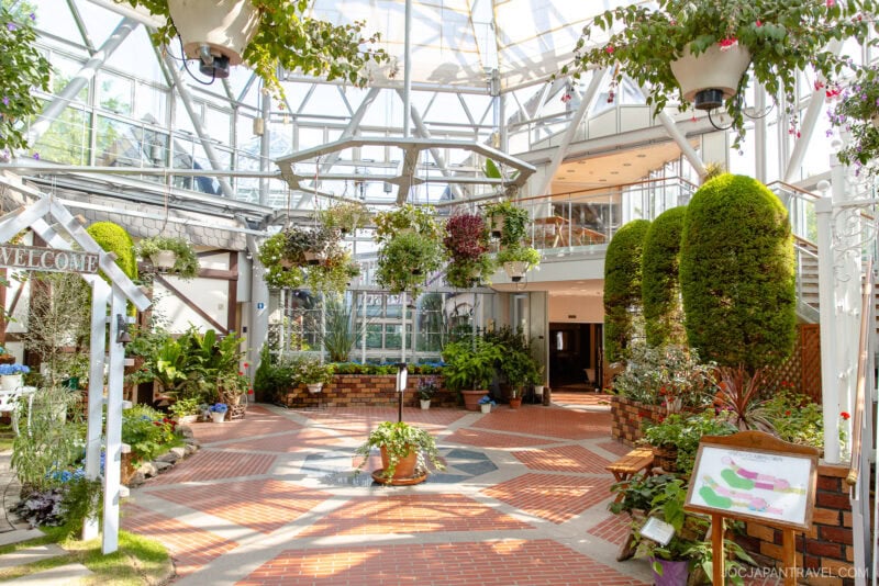 A bright indoor garden with glass walls and ceiling, inspired by the Nunobiki Ropeway and Herb Gardens, is filled with lush green plants, hanging baskets, and potted flowers. A Welcome sign stands to the left as sunlight streams in from above.