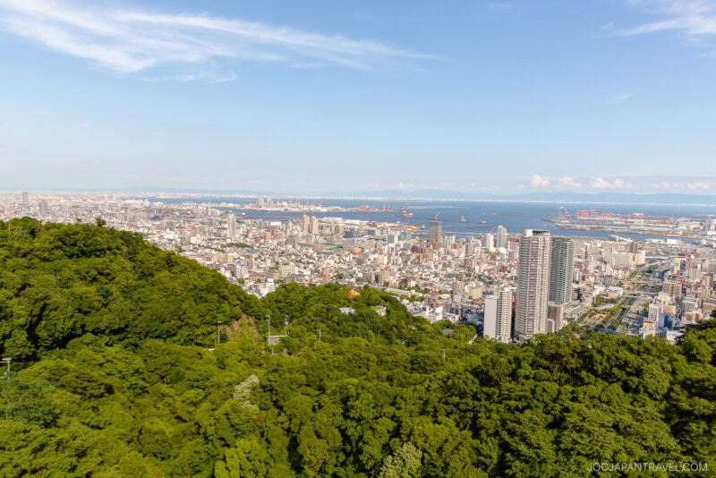 A panoramic view of the city by the sea, seen from a high vantage point near dense green trees under a blue sky with scattered clouds, showcasing the Nunobiki Ropeway and Herb Gardens in the lush foreground.