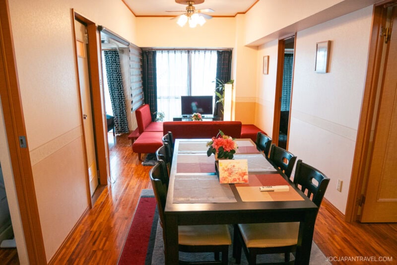 A dining room with a table set for six, a flower centerpiece, and placemats. Beyond is a cozy living area with a red sofa, TV, and large window. This bright Okinawa AirBnb features wooden floors&mdash;and easy access to rental car options.
