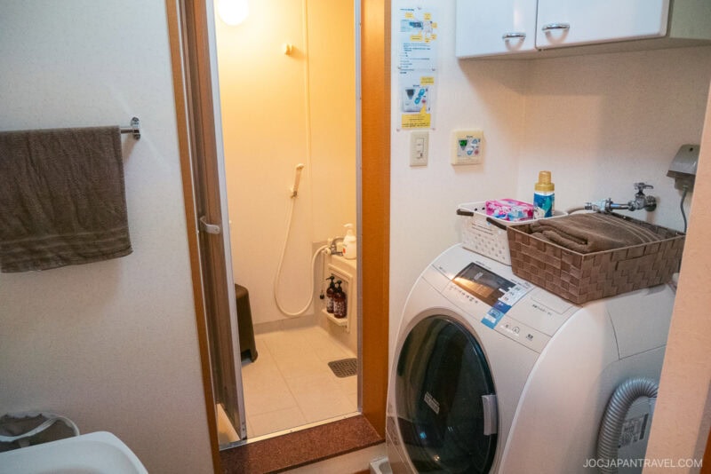 A compact Japanese bathroom in an Okinawa AirBnb, featuring a washing machine, sink, towel rack, and storage baskets. An open door reveals a small shower area with a stool and toiletries—a clean, organized space for your rental car adventures.