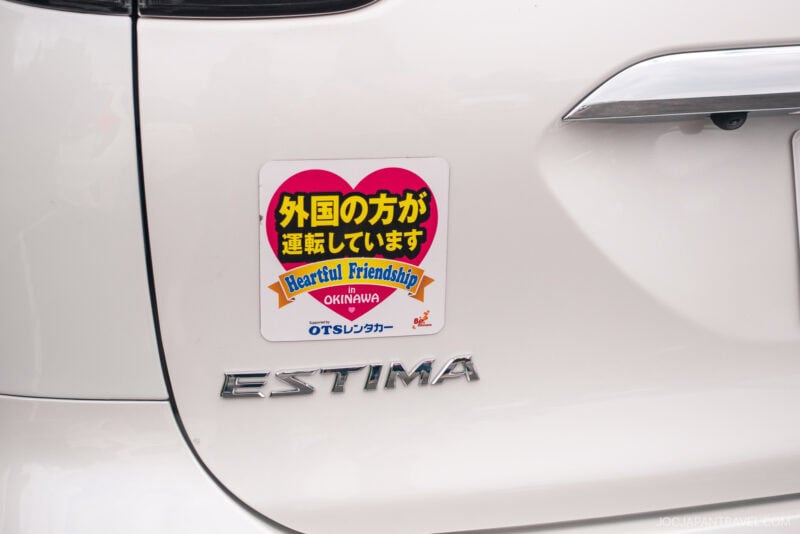 A sticker on a white car reads, 外国の方が運転しています, with Heartful Friendship, Okinawa, and Okinawa AirBnb and Rental Car in English below—indicating a foreign driver. The car badge says ESTIMA.