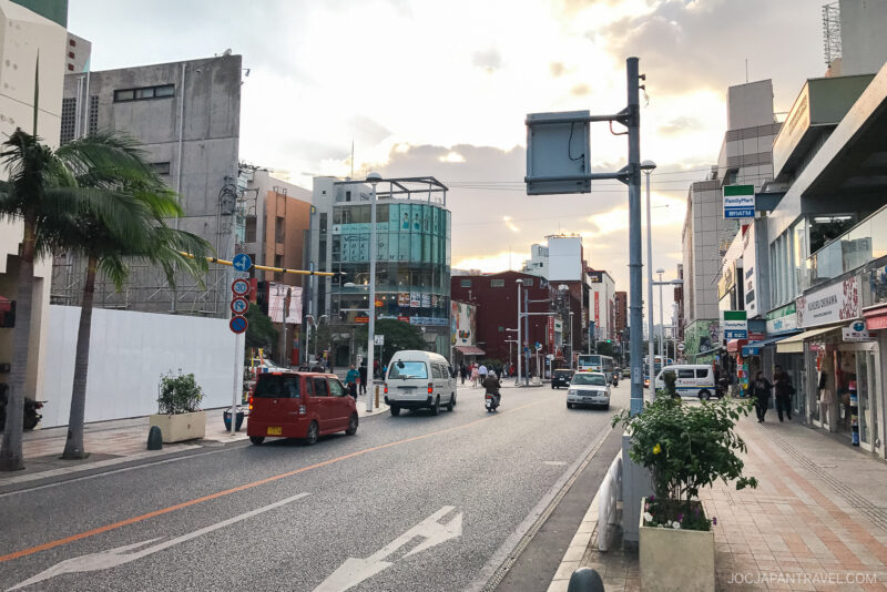 A city street scene in Okinawa with cars, a van, and a motorcycle—perfect for exploring with an Airbnb stay and rental car. Shops and buildings line both sides, pedestrians stroll the sidewalks, and palm trees sway under partly cloudy skies.