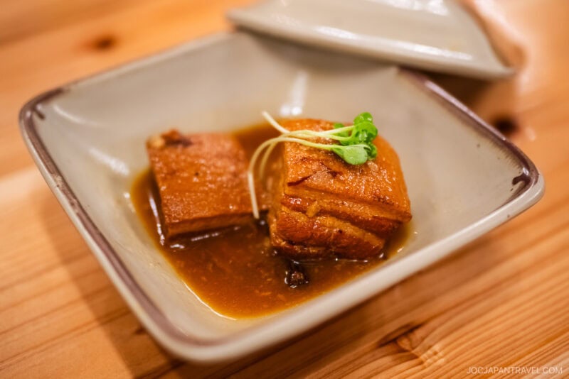 A square dish with two pieces of braised pork belly in brown sauce, garnished with microgreens, showcases a classic Okinawa Food specialty, beautifully presented on a wooden table.