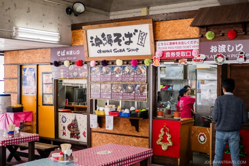 A Japanese food stall decorated with festive ornaments serves Okinawa soba soup and other Okinawa food. Menu pictures are displayed, and signs in Japanese and English indicate Soba Pickup and Please Press Food Ticket Here. Tables are in front.