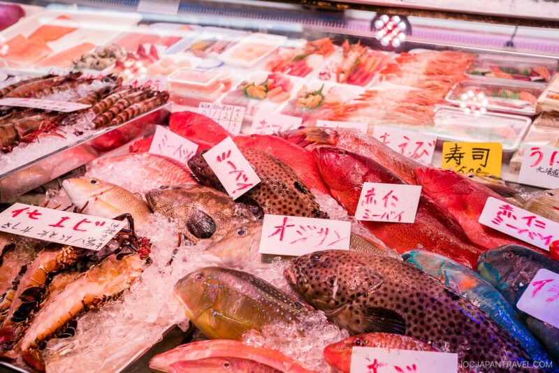 A seafood market display features various fresh fish, shrimp, and other Okinawa food specialties laid on ice, with handwritten Japanese labels identifying each item. The vibrant colors highlight the freshness of the seafood.