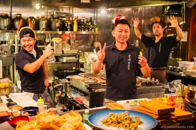 Three cheerful chefs pose playfully in a vibrant, bustling Japanese kitchen, surrounded by Okinawa food specialties and fresh ingredients, one flashing a peace sign and another with hands up like antlers at the lively counter.