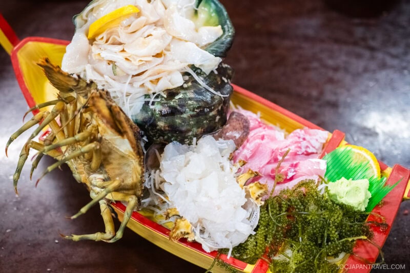 A colorful wooden boat platter filled with assorted sashimi, showcasing Okinawa food delights like raw fish, sea grapes, shellfish, and garnishes, all artfully arranged for a vibrant presentation.