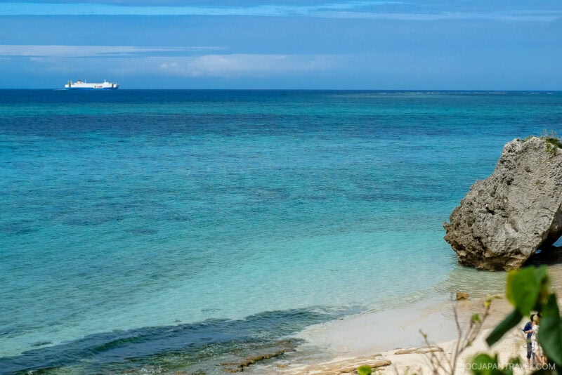 A couple in wedding attire stands on a sandy beach near a large rock at Okinawa Ocean Expo Park, overlooking clear blue-green ocean waters, with a cargo ship visible on the horizon under a vivid blue sky.
