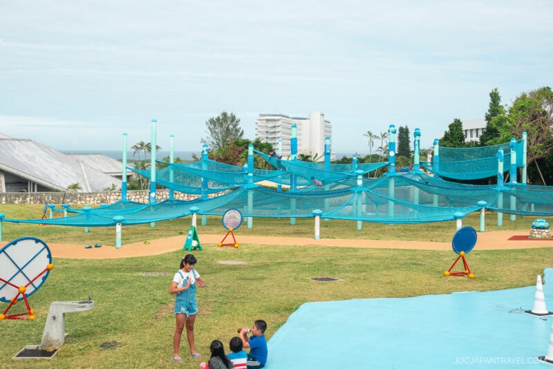 Children play on the grass near a blue net climbing structure in an outdoor playground at Okinawa Ocean Expo Park, with modern buildings and trees in the background under a partly cloudy sky.