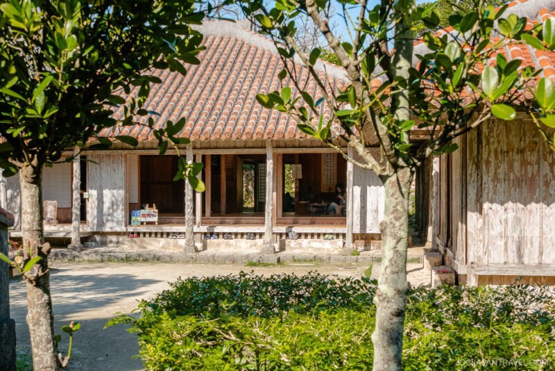 A traditional Japanese wooden house with a tiled roof, surrounded by green bushes and trees, sits under bright sunlight near Okinawa Ocean Expo Park. The structure features open walls and a shaded porch area.