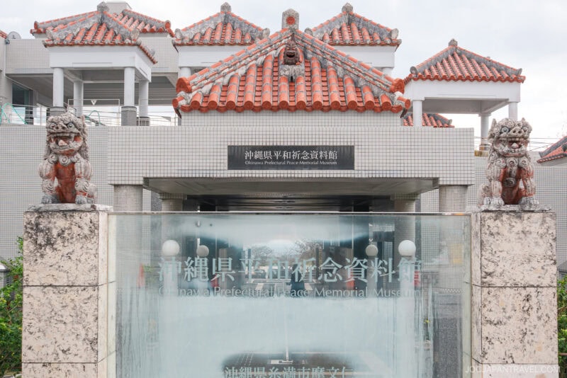 Entrance to the Okinawa Prefectural Peace Memorial Museum in Okinawa Prefectural Peace Memorial Park, featuring traditional red-tiled roofs, two Shisa lion statues, and glass signage with Japanese and English text.