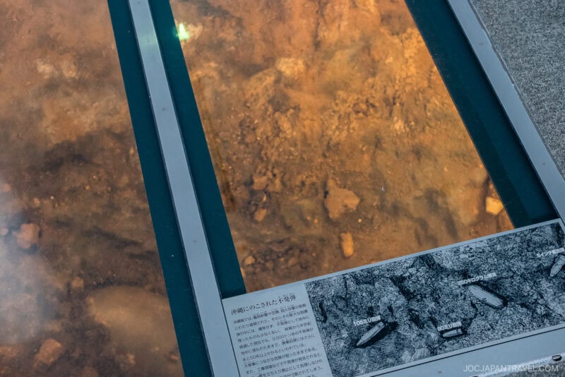 A glass-covered display reveals ancient ruins or artifacts embedded in soil, accompanied by an informational panel with text and a labeled diagram at Okinawa Prefectural Peace Memorial Park.