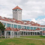A modern building with traditional red-tiled rooftops and a central tower, reminiscent of Okinawa Prefectural Peace Memorial Park, set against a partly cloudy sky, with a grassy lawn in the foreground.