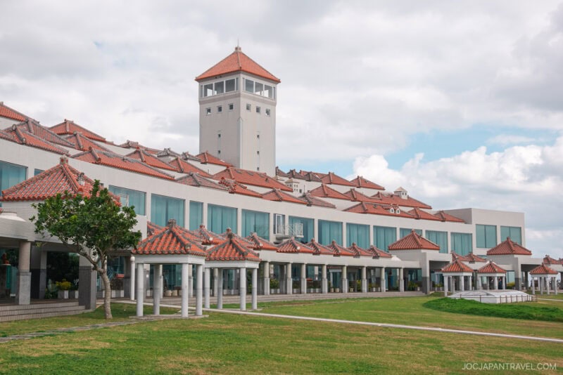 A modern building with traditional red-tiled rooftops and a central tower, reminiscent of Okinawa Prefectural Peace Memorial Park, set against a partly cloudy sky, with a grassy lawn in the foreground.