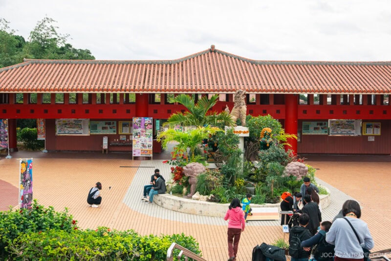 A group of people gather in a brick-paved courtyard with a circular garden and palm trees at Okinawa World, in front of a red building with a tiled roof and informational boards on the wall.