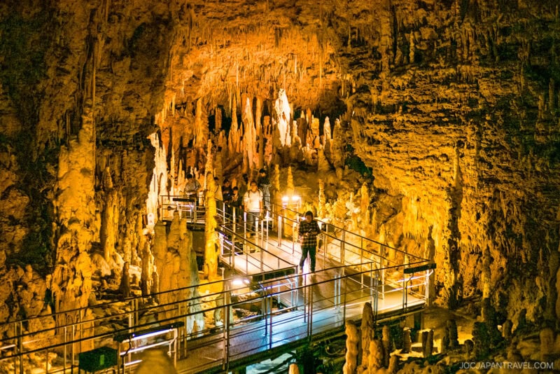 A person stands on a metal walkway surrounded by illuminated stalactites and stalagmites inside the golden-lit Gyokusendo Cave at Okinawa World.